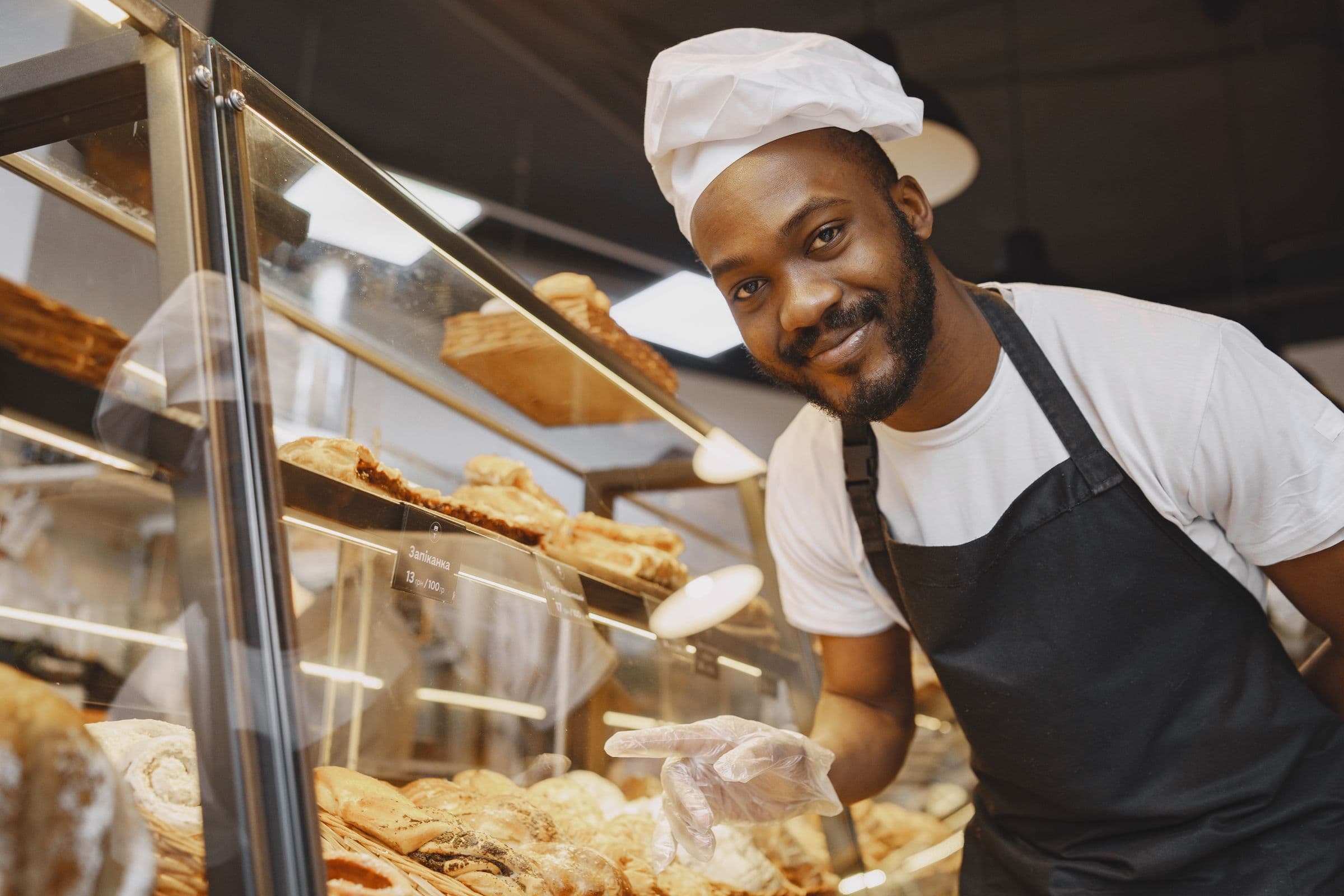 A baker in an apron behind a bakery counter, with trays of fresh pastries in the lit display case beside him.